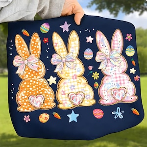 Peut inclure: Une couverture bleu marine sur le thème de Pâques avec trois illustrations de lapins. Les lapins ont une fourrure à motifs et des nœuds, entourés d'œufs de Pâques, de carottes, d'étoiles et de fleurs. La couverture est tenue par une personne.