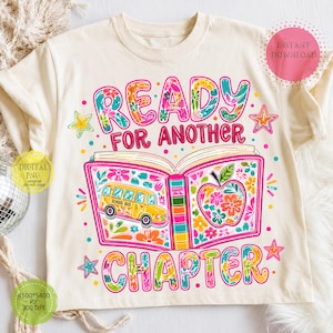 May include: A cream-colored t-shirt featuring a graphic of an open book with floral patterns, a yellow school bus, and an apple. The text reads READY FOR ANOTHER CHAPTER. The design includes colorful stars and dots, with digital download labels.
