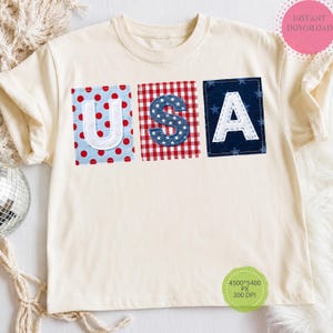 May include: Cream-colored t-shirt with the letters "USA" appliqued on the front. The letters are on patches with red polka dots, red and white gingham, and blue with white stars. The letter outlines are white.