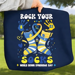 May include: A navy blue tote bag with the words "ROCK YOUR SOCKS" in white and yellow. The design includes two cartoon socks, a ribbon, handprints, hearts, and the text "WORLD DOWN SYNDROME DAY".