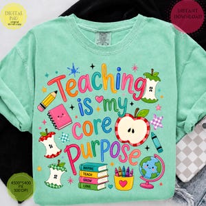 May include: A mint green t-shirt featuring a colorful graphic with the text Teaching is my core PURPOSE. The design includes illustrations of a pencil, a pink notebook, apple cores, a stack of books labeled Inspire, Teach, Grow, Love, a cup of crayons, and a globe.