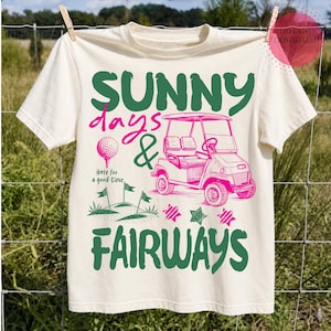 May include: A cream-coloured t-shirt with a golf-themed design. The graphic includes the words "SUNNY days & FAIRWAYS" in green and pink, a golf cart, golf ball, and flags. The text "Here for a good time" is also present.