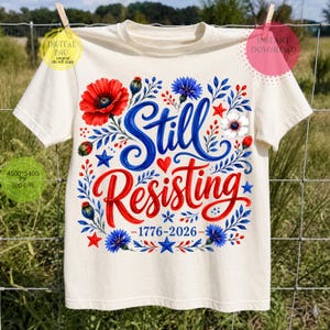 May include: A cream-colored t-shirt featuring the blue text Still and red text Resisting, surrounded by red poppies, blue cornflowers, small red hearts, and blue stars. The text 1776-2026 is printed in blue below the main graphic design.