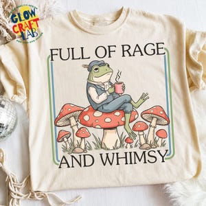 以下が含まれることがあります： クリーム色のTシャツ。帽子とベストを着たカエルがキノコに座り、マグカップを持っています。「FULL OF RAGE AND WHIMSY」の文字が緑と青のフレームで囲まれています。追加のキノコのイラストも含まれています。