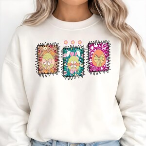 Può includere: Felpa bianca con tre disegni di uova di Pasqua colorate. Ogni uovo è incorniciato da un bordo nero a zig-zag e decorato con dettagli floreali e fiocchi. I disegni includono colori pastello e un uovo con orecchie da coniglio.