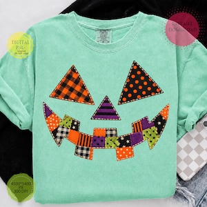 May include: A mint green t-shirt featuring a large jack-o'-lantern face graphic made of various patterned fabric patches. The eyes are orange plaid and orange polka dot triangles, the nose is a purple striped triangle, and the mouth is a patchwork grin.