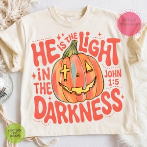 May include: A cream-colored t-shirt featuring a graphic of an orange pumpkin with a cross-shaped eye and a wide smile. The text reads "HE IS THE LIGHT IN THE DARKNESS" and "JOHN 1:5" in bold red lettering. The design includes small star accents.