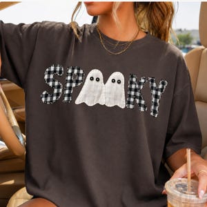May include: A dark gray short-sleeve t-shirt featuring the word SPOOKY in a black and white buffalo plaid pattern. The two letter O's are replaced by white ghost shapes with black button eyes, creating a graphic design across the chest area.