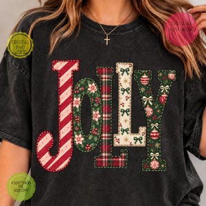 May include: A charcoal gray t-shirt featuring the word JOLLY in large letters. The J has red and white candy cane stripes, the O has a green poinsettia pattern, the L has a red and green plaid design, the L has a cream background with green bows, and the Y has a green background with red ornaments.