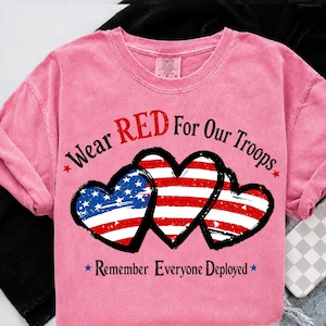 May include: Pink t-shirt with the text "Wear RED For Our Troops" and "Remember Everyone Deployed." The design features three hearts with the American flag design. The shirt is a crew neck.