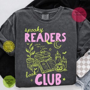 May include: A charcoal gray t-shirt featuring the text "spooky READERS book CLUB" in pink and lime green. The graphic includes a stack of books, a potted plant, a coffee mug, two candles, a jack-o'-lantern, a crescent moon, and bats.
