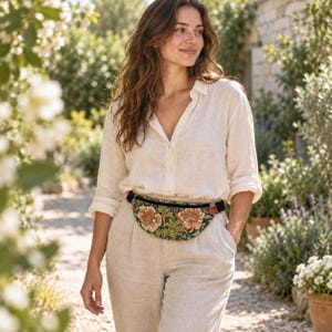May include: A waist bag featuring a dark green fabric base with a repeating floral pattern of large orange and cream-colored blossoms and green leaves. The bag has a black zipper closure and an adjustable black strap worn around the waist.