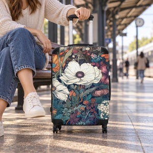 May include: A rolling suitcase featuring a dark, intricate botanical print with large white poppy-like flowers, deep blue foliage, and accents of orange and purple. The luggage has a black frame, a retractable silver handle, and four wheels.