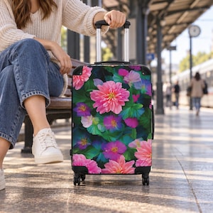 May include: A rolling suitcase featuring a vibrant floral pattern with large pink dahlias and smaller purple and blue flowers set against a dark black background. The luggage has a retractable silver handle and four black wheels for mobility.