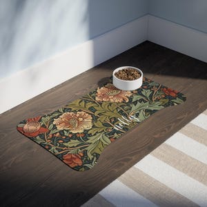 May include: A bone-shaped pet feeding mat featuring a dark green background with a dense pattern of orange and yellow flowers and green leaves. The name "Nelly" is printed in white cursive script on the mat, which holds a white bowl of dry food.