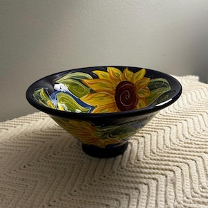 May include: A deep, conical ceramic bowl featuring a dark navy blue background adorned with hand-painted yellow sunflowers and green leaves. The interior and exterior surfaces display a glossy finish with vibrant floral motifs encircling the bowl.