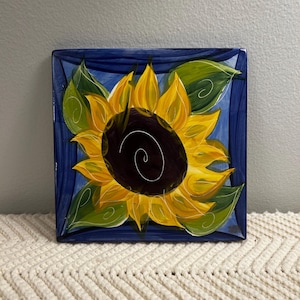 May include: A square ceramic plate featuring a hand-painted sunflower with bright yellow petals and a dark brown center. The flower is surrounded by green leaves against a deep blue background, with a white spiral detail on the center of the flower.