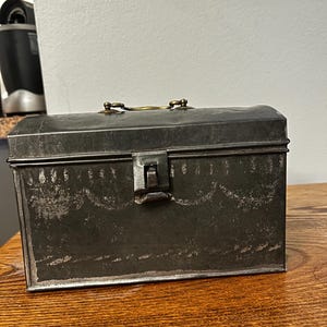 May include: A weathered, rectangular metal box with a hinged lid and a small handle. The box is a dark, muted gray color with visible wear and tear, suggesting age. The latch is a simple, functional design. The box sits on a wooden surface.