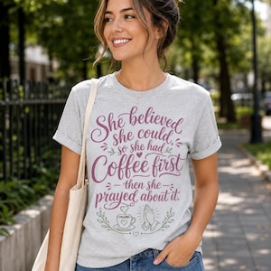 May include: A heather gray short-sleeve t-shirt featuring the maroon text "She believed she could, so she had Coffee first then she prayed about it." The design includes a small coffee cup and praying hands graphic with green leaf accents.