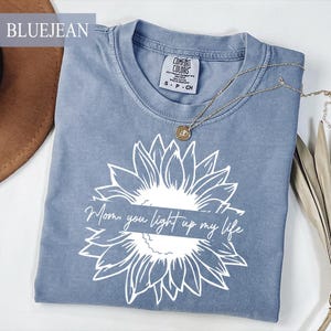 Puede incluir: Camiseta azul vaquero con un diseño de girasol blanco y el texto "Mom, you light up my life." Un collar dorado con un colgante descansa sobre la camiseta. La camiseta es de la marca Comfort Colors.