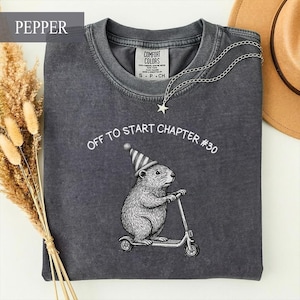 May include: A charcoal gray crewneck t-shirt featuring a white graphic of a groundhog wearing a party hat and riding a scooter. The text above the graphic reads OFF TO START CHAPTER #30. A silver chain necklace with a star charm rests on the collar.