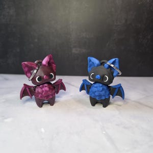 May include: Two bat-shaped keychains with metal rings. One keychain is dark magenta with a textured body and smooth wings. The other keychain is black and blue with a textured body and smooth wings. Both have large white eyes and pointed ears.