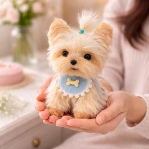 May include: A plush Yorkshire Terrier puppy with tan and cream fur, wearing a blue bib with a bone-shaped design. The puppy has large, dark eyes and a small ponytail secured with a blue band. The puppy is being held in someone's hands.