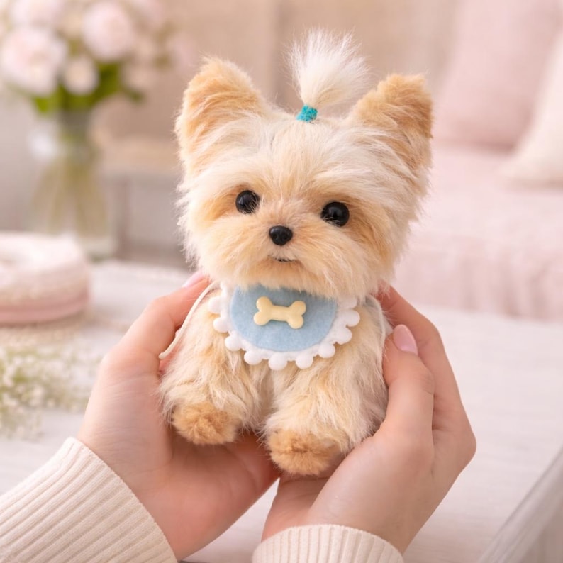 May include: A plush Yorkshire Terrier puppy with tan and cream fur, wearing a light blue bib with a bone-shaped design. The puppy has a small ponytail and is held in someone's hands.