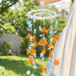 May include: A decorative mobile featuring orange fish and blue star-shaped ornaments suspended from a white, beaded ring. The mobile is held up against a blurred green background, suggesting an outdoor setting. The fish and stars are attached with white ribbons.