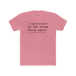 May include: A dusty pink short-sleeve t-shirt featuring black typewriter-style text that reads: i hyperfocused on the wrong thing again * not for the first time. The shirt has a crew neckline and a standard fit, made of a solid-colored fabric.