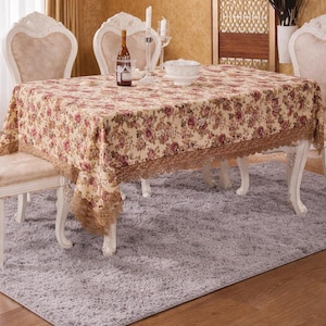 May include: A dining room table set with a floral tablecloth, white chairs, and a gray rug. The tablecloth has a beige background with pink and red floral patterns and a lace trim. A bottle of wine, candles, and a bowl are on the table.