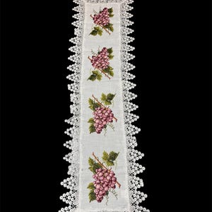 May include: A white rectangular table runner with a detailed lace border. The runner features embroidered clusters of purple grapes with green leaves and brown stems. The lace border adds a delicate touch to the design.