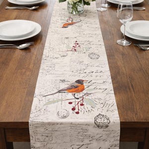 May include: A rectangular table runner with a bird and berry design. The runner is cream-colored with a vintage script pattern and features an orange and gray bird perched on a branch with red berries. The runner is placed on a wooden table.