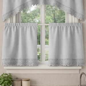 May include: Light gray kitchen curtains with a lace trim. The set includes a valance and two shorter panels. The curtains are hanging in front of a window with a view of trees. The kitchen counter is visible in the foreground.