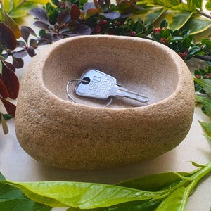 May include: A small, beige stone bowl containing a key. The key is silver and marked with "SI". The bowl is set amongst green and yellow leaves and branches with small red berries.