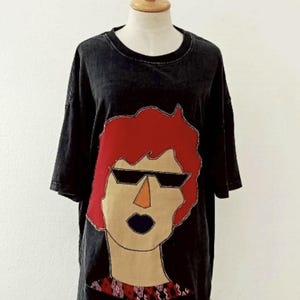 Puede incluir: Camiseta negra con un diseño gráfico de una persona con pelo rojo, gafas de sol y un labio azul oscuro. El diseño está en la parte delantera de la camiseta. La camiseta se muestra en un maniquí.