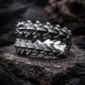 Peut inclure: Une bague argentée avec un design en forme d'épine dorsale, posée sur une surface sombre et texturée. Les détails complexes de la bague ressortent sur un fond flou, avec un crâne et des plumes ajoutant à l'esthétique gothique.