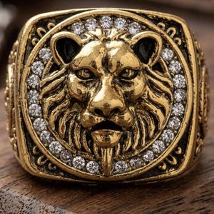 Peut inclure: Bague dorée ornée d'une tête de lion détaillée. Le visage du lion est entouré de petites pierres précieuses claires. La bague est de forme rectangulaire avec des détails ornés.