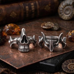 Peut inclure: Deux bagues argentées en forme de crâne avec des détails squelettiques. Les bagues ont une esthétique gothique ou motard, avec l'accent mis sur les dents et les orbites du crâne. Elles sont présentées sur une surface sombre et texturée.