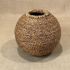 May include: A round, woven basket with a wide opening. The basket is made of natural-colored fibers, tightly woven in a spiral pattern. The interior of the basket is dark. The basket sits on a neutral-toned surface.