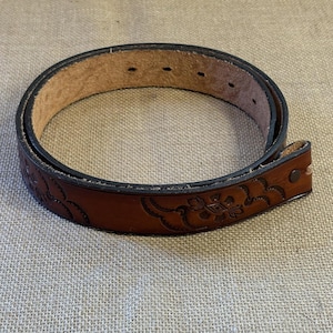 Puede incluir: Un cinturón de cuero marrón con un diseño floral en relieve. El cinturón tiene un borde negro y varios agujeros para ajustar. El extremo de la hebilla está asegurado con un remache metálico. El cinturón mide aproximadamente 3,8 cm de ancho.