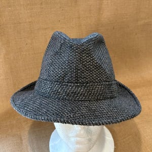 May include: A gray fedora hat with a textured weave pattern. The hat features a structured crown, a wide brim, and a band around the base of the crown. The hat is displayed on a white mannequin head.