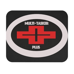 Op de afbeelding: Een rechthoekige muismat met afgeronde hoeken, een zwarte achtergrond en een grote witte ovale omlijning. Binnenin het ovaal staat een rode kruisvormige afbeelding met een zwarte plus-uitsparing, met de tekst MULTI-TASKER erboven en PLUS eronder.