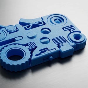 May include: A blue, abstract, geometric object with various cut-out shapes, including circles, a gear, and curved lines. The object is made of a solid material and sits on a metallic surface.