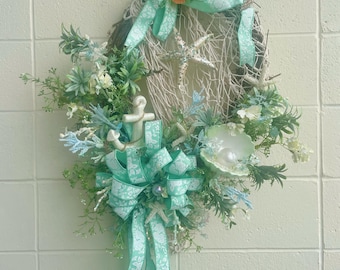 May include: An oval-shaped coastal wreath with a woven base, adorned with faux greenery, seashells, and starfish. The wreath features a large bow and ribbons in shades of seafoam green and white. The text "JLowsDesigns" is visible.