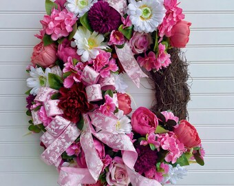 May include: A floral wreath with a variety of pink, white, and burgundy flowers, accented with pink ribbons. The wreath is made of a natural grapevine base and is displayed against a white background. The text "JlowsDesigns" is visible.