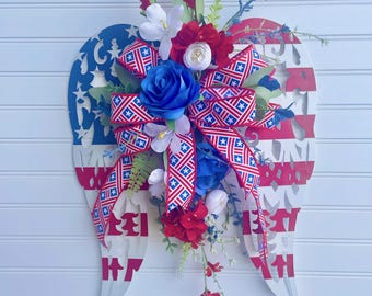 May include: A patriotic wall hanging shaped like angel wings, painted in red, white, and blue. The wings are adorned with a floral arrangement featuring red, white, and blue flowers and a star-patterned ribbon. The text "HOME" is visible on the wings.