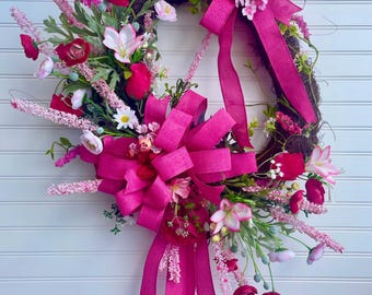 May include: A vibrant floral wreath with a brown base, adorned with pink and red flowers, green foliage, and large fuchsia bows. The wreath is displayed against a white background. The text "JlowsDesigns" is visible in the bottom right corner.