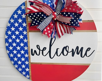 May include: A round wooden welcome sign with a patriotic theme. The sign features a red, white, and blue color scheme with a large bow at the top. The word "welcome" is written in black script across the center.