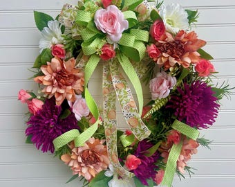 May include: A vibrant floral wreath featuring a variety of colorful flowers, including pink roses, orange dahlias, and deep purple blooms. The wreath is adorned with green ribbons and a patterned ribbon, creating a cheerful and decorative accent.